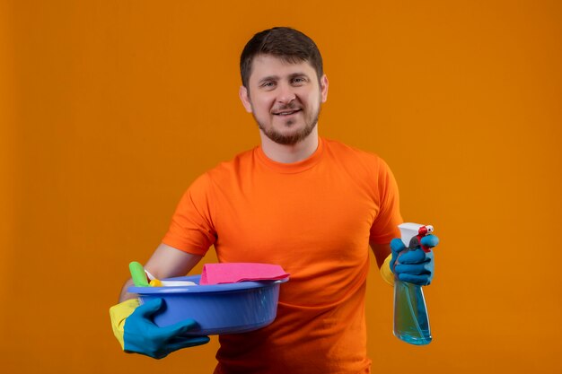 Jeune homme portant un t-shirt orange tenant le bassin avec des outils de nettoyage et un spray de nettoyage souriant regardant la caméra positive et heureux prêt à nettoyer debout sur fond orange 2