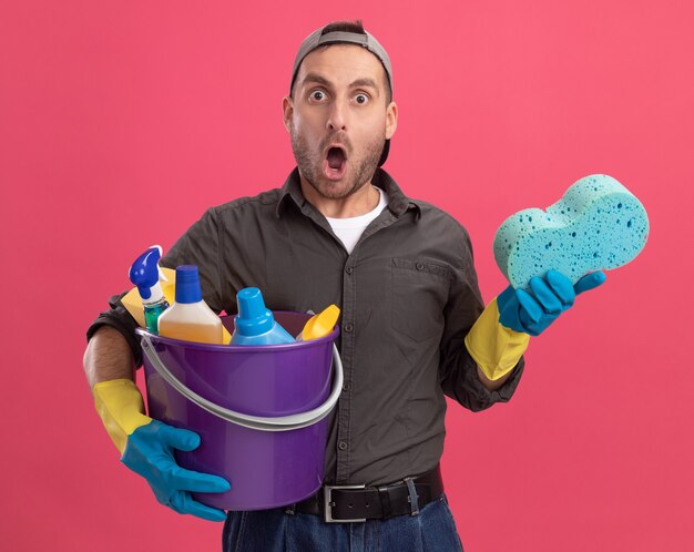 Jeune homme portant des vêtements décontractés et une casquette de gants en caoutchouc tenant un seau avec des outils de nettoyage et une éponge à la surprise et surpris debout sur le mur rose