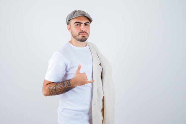 Jeune homme posant à l'avant avec une veste sur l'épaule et montrant un geste d'appel en t-shirt blanc, veste et casquette grise et l'air sérieux