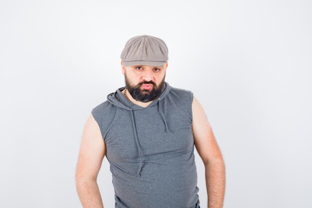 Jeune homme posant debout dans un sweat à capuche sans manches, une casquette et l'air confiant. vue de face.