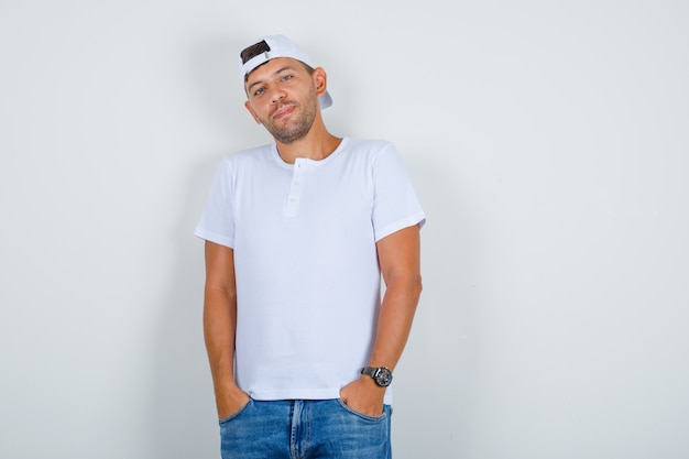 Jeune homme posant avec les mains dans les poches en t-shirt blanc, casquette, jeans et regardant beau, vue de face