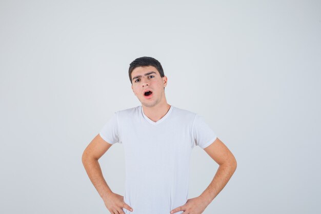 Jeune homme posant avec les mains sur la taille tout en ouvrant la bouche en t-shirt et regardant perplexe, vue de face.