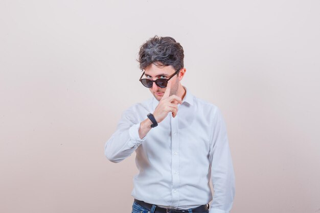 Jeune homme posant tout en regardant par-dessus des lunettes en chemise blanche, jeans et élégant