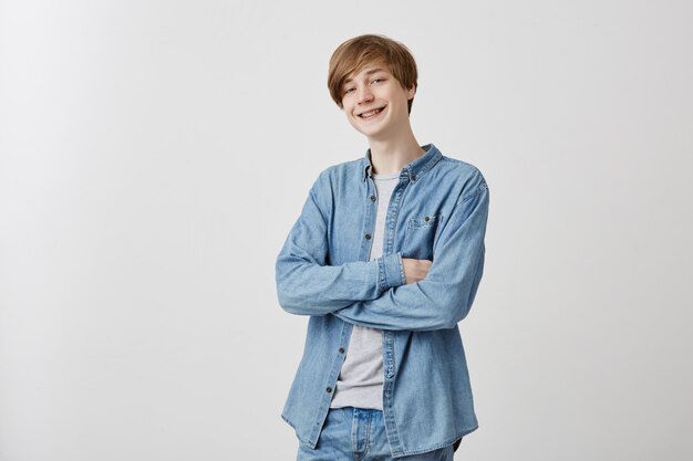 Jeune homme positif en chemise en jean aux cheveux blonds et aux yeux bleus, timide, souriant nerveusement en position fermée, gardant les bras croisés tout en parlant à la fille, il aime