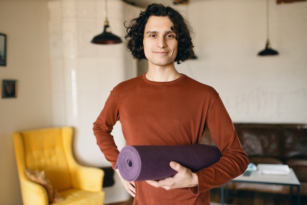 Jeune homme positif dans des vêtements décontractés tenant un rouleau de tapis de yoga sous son bras va pratiquer à l'intérieur, heureux de rester à la maison pendant la quarantaine, passant plus de temps sur le développement personnel et une activité saine