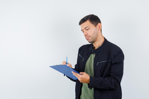 Jeune homme prenant des notes sur le presse-papiers en t-shirt, veste et l'air occupé. vue de face.