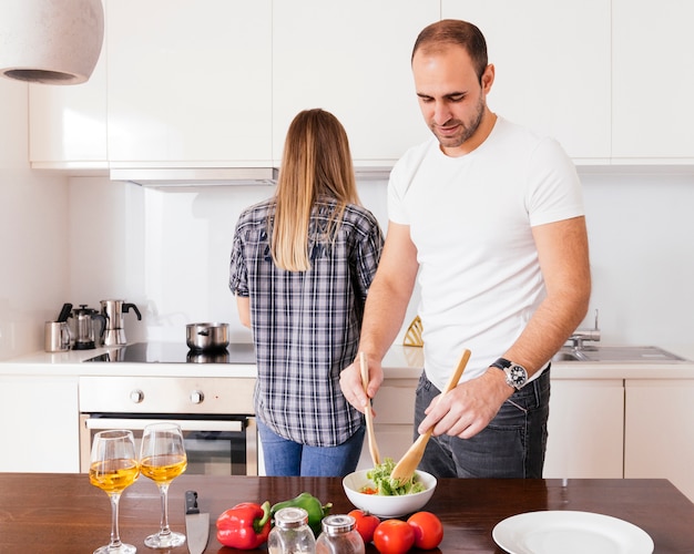 Jeune homme préparant la salade et sa femme debout derrière lui, préparant un repas dans la cuisine