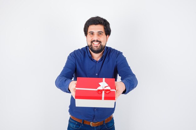 Jeune homme présentant une boîte-cadeau avec les deux mains en chemise bleue et en jean et l'air optimiste. vue de face.