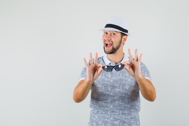 Jeune homme présentant des lunettes de soleil en t-shirt, chapeau et à la vue positive, de face.