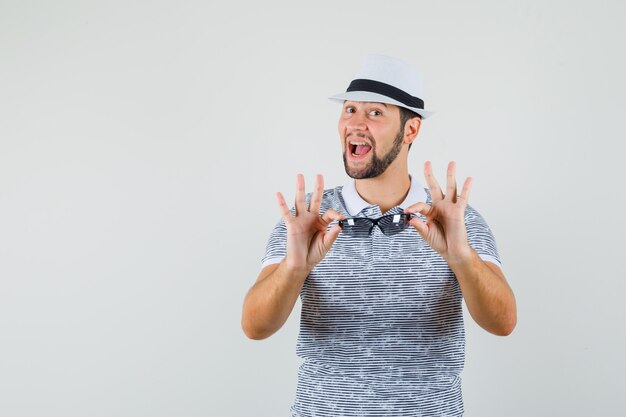 Jeune homme présentant des lunettes de soleil en t-shirt, chapeau et à la vue positive, de face.