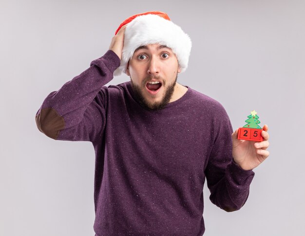 Jeune homme en pull violet et bonnet de Noel montrant des cubes avec la date du nouvel an regardant la caméra étonné et surpris debout sur fond blanc