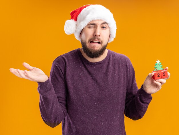 Jeune homme en pull violet et bonnet de Noel montrant des cubes de jouet avec numéro vingt-cinq confus et mécontent en haussant les épaules debout sur le mur orange