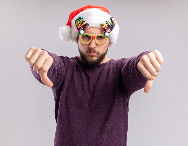 Jeune homme en pull violet et bonnet de Noel portant des lunettes drôles regardant la caméra avec un visage en colère montrant les pouces vers le bas debout sur fond blanc