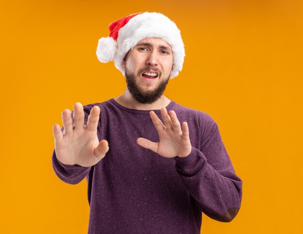 Jeune homme en pull violet et bonnet de Noel regardant la caméra inquiète faisant le geste de défense tenant la main en disant ne pas se rapprocher debout sur fond orange