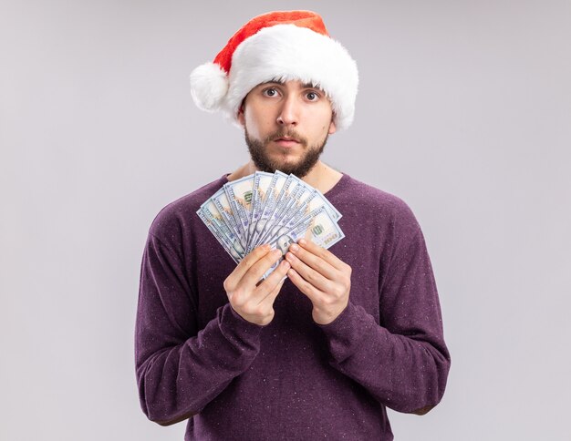 Jeune homme en pull violet et bonnet de noel tenant de l'argent en regardant la caméra avec un visage sérieux debout sur fond blanc