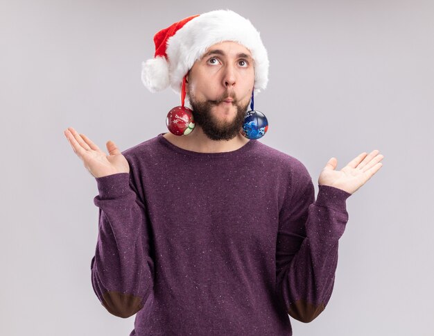 Jeune homme en pull violet et bonnet de noel tenant des boules de Noël à la confusion avec les bras levés debout sur fond blanc