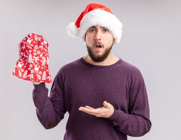 Jeune homme en pull violet et bonnet de Noel tenant un sac rouge avec des cadeaux présentant avec bras à la confusion debout sur fond blanc