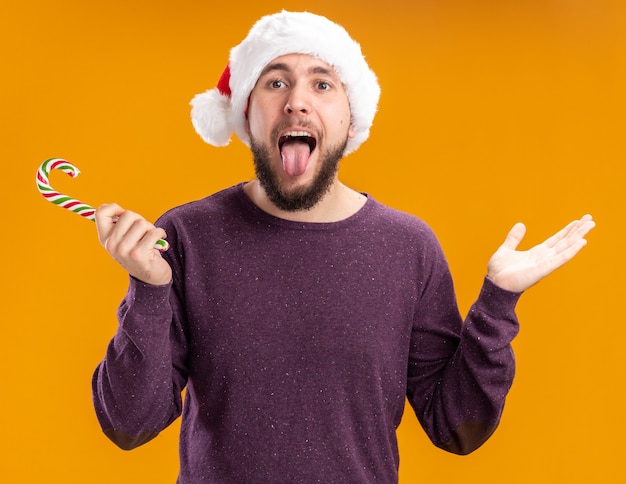 Jeune homme en pull violet et santa hat holding candy cane looking at camera sticking out tongue heureux et gai debout sur fond orange