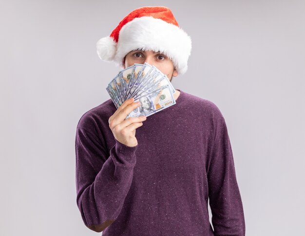 Jeune homme en pull violet et santa hat holding cash couvrant le visage avec de l'argent regardant la caméra avec un visage sérieux debout sur fond blanc