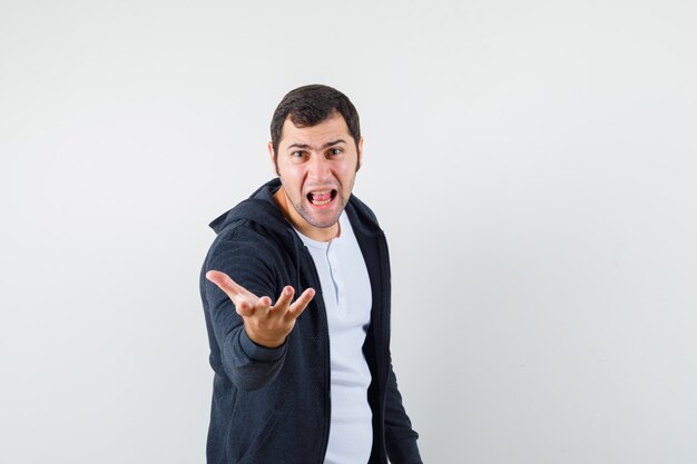 Jeune homme qui s'étend la main dans le geste d'interrogation en t-shirt, veste et à la colère. vue de face.