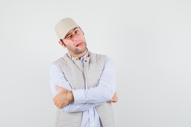 Jeune homme qui sort la langue tout en gardant les bras croisés en chemise, veste sans manches, casquette et à la folie. vue de face.