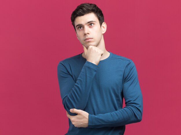 Un jeune homme de race blanche brune réfléchie met la main sur le menton en regardant le côté isolé sur un mur rose avec un espace de copie