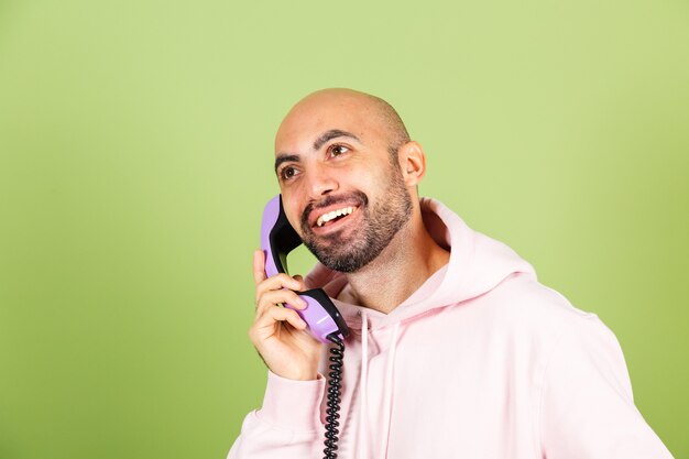 Jeune homme de race blanche chauve en sweat à capuche rose isolé, maintenez le téléphone fixe avec heureux visage étonné excité