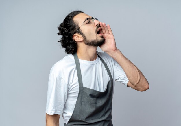 Jeune homme de race blanche coiffeur portant des lunettes et bande de cheveux ondulés en uniforme regardant côté mettant la main près de la bouche appelant à quelqu'un isolé sur fond blanc avec espace de copie