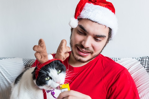 Jeune homme de race blanche dans un chapeau de père Noël nourrir son chat blanc et noir dans des cornes d'élan