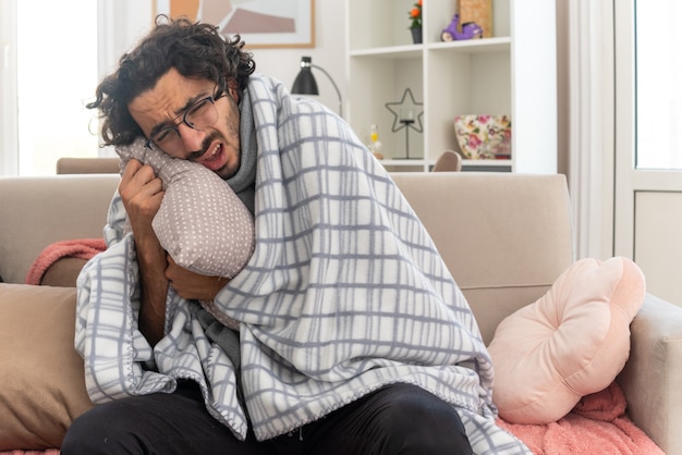 Jeune homme de race blanche malade endolori dans des lunettes optiques enveloppées dans un plaid avec une écharpe autour du cou serrant l'oreiller assis sur un canapé dans le salon