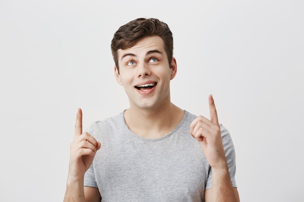 Jeune homme de race blanche portant un t-shirt gris pointant vers l'espace de copie avec les index à la recherche avec un large sourire vers le haut démontrant quelque chose au fond du studio blanc. Concept de langage corporel