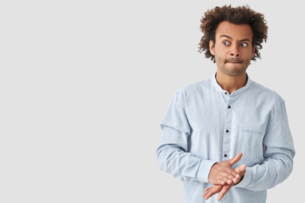 Un jeune homme de race mixte perplexe mord les lèvres, a les cheveux croquants, garde les mains jointes, regarde de côté avec confusion, vêtu d'une chemise à la mode, pose contre un mur blanc