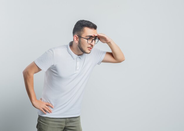 Jeune homme à la recherche de loin avec la main sur la tête en t-shirt blanc, pantalon et à concentré