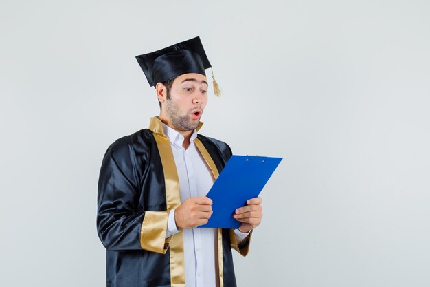 Jeune homme à la recherche de notes sur le presse-papiers en uniforme d'études supérieures et à la surprise, vue de face.