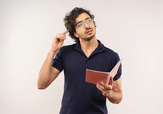 Jeune homme réfléchi en chemise noire avec des lunettes optiques tient le cahier et met un stylo sur la tête isolé sur un mur blanc