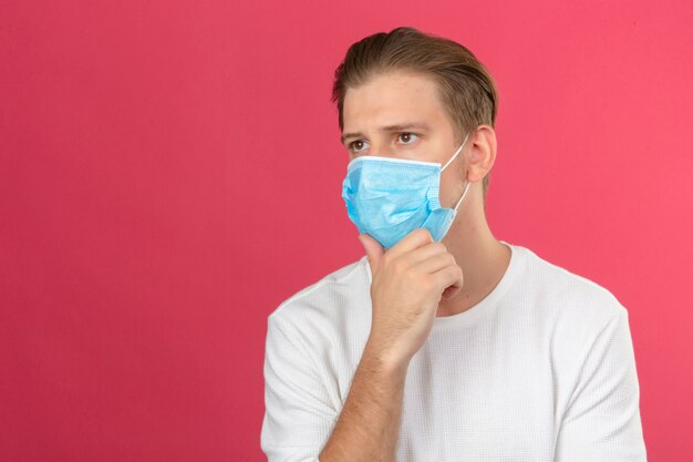 Jeune homme réfléchi dans un masque de protection médicale touchant le menton et la réflexion sur fond rose isolé