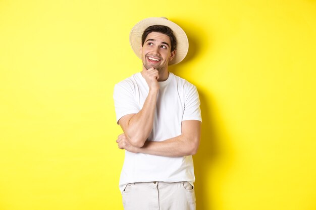 Jeune homme réfléchi touriste imageant quelque chose, regardant le coin supérieur gauche et souriant, pensant et se tenant sur fond jaune