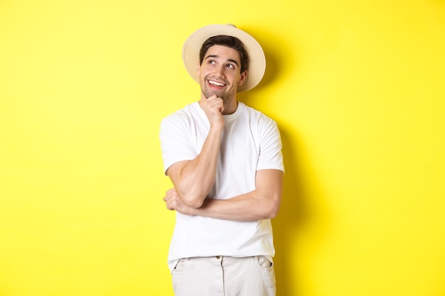 Jeune homme réfléchi touriste imageant quelque chose, regardant le coin supérieur gauche et souriant, pensant et se tenant sur fond jaune