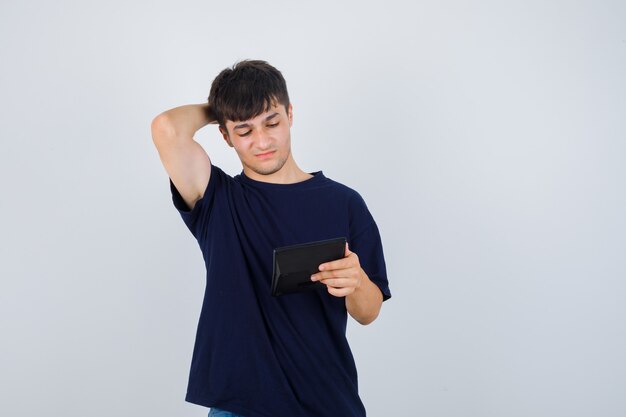 Jeune homme regardant la calculatrice, gardant la main derrière la tête en t-shirt noir et regardant perplexe, vue de face.