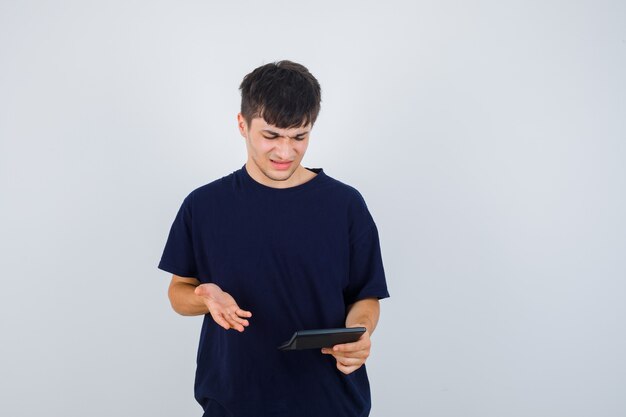 Jeune homme regardant la calculatrice en t-shirt noir et à la confusion. vue de face.