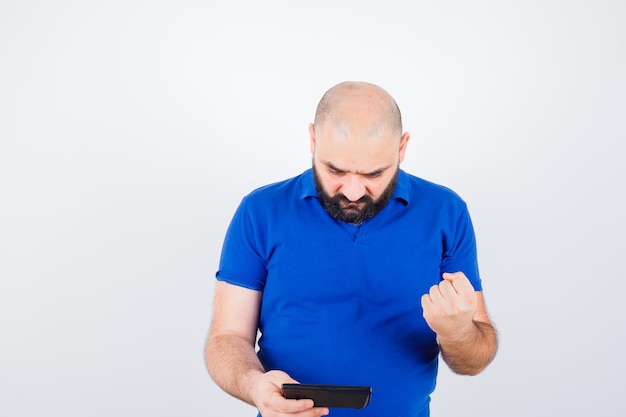 Jeune homme regardant la calculatrice tout en levant le poing en chemise bleue et en ayant l'air réussi. vue de face.