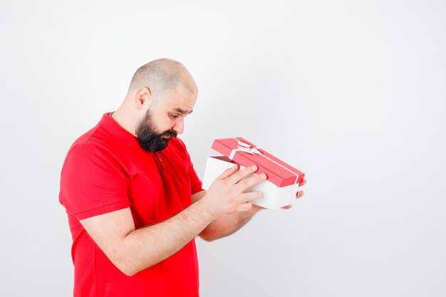 Jeune homme regardant dans une boîte-cadeau en t-shirt rouge et se demandait. vue de face.