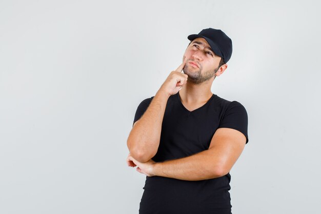 Jeune homme regardant avec le doigt sur la joue en t-shirt noir, casquette et à la pensif