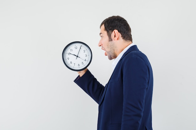 Jeune homme regardant horloge murale en chemise et veste et à la surprise.