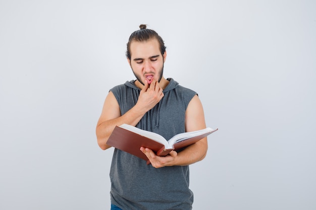 Jeune homme regardant livre tout en tenant la main sur la bouche en sweat à capuche sans manches et à la vue réfléchie, de face.