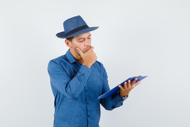 Jeune homme regardant par-dessus les notes sur le presse-papiers en chemise bleue, chapeau et pensif. vue de face.