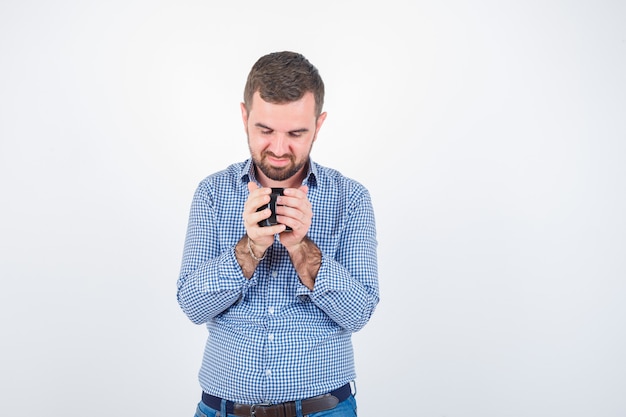 Photo gratuite jeune homme regardant la tasse en chemise, jeans et regardant heureux, vue de face.