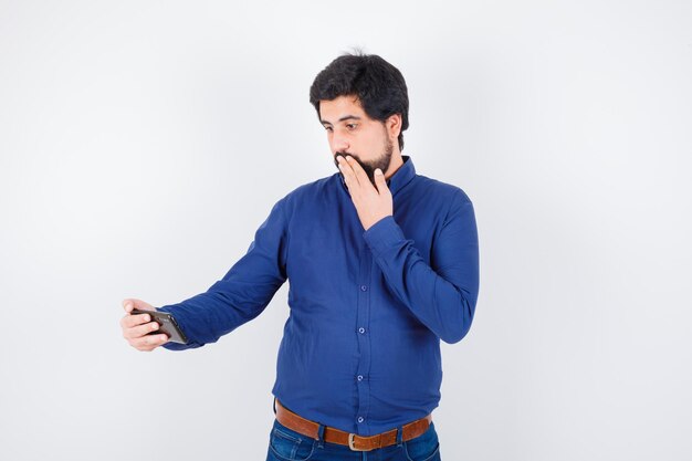 Jeune homme regardant le téléphone avec la main sur la bouche en chemise bleu royal et l'air terrifié, vue de face.