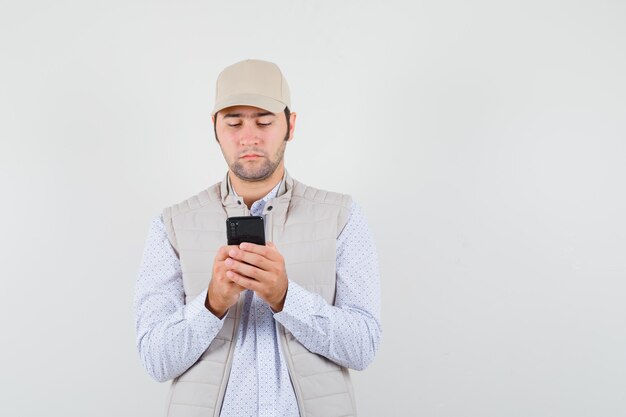 Jeune homme regardant un téléphone mobile en veste beige et casquette et à la recherche focalisée, vue de face.