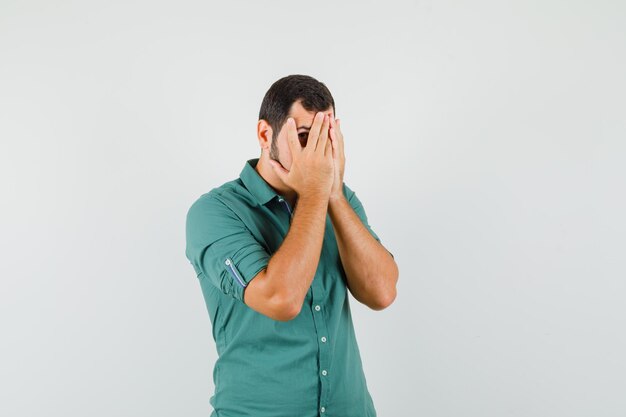 Jeune homme regardant à travers les doigts en chemise verte et l'air inquiet, vue de face.